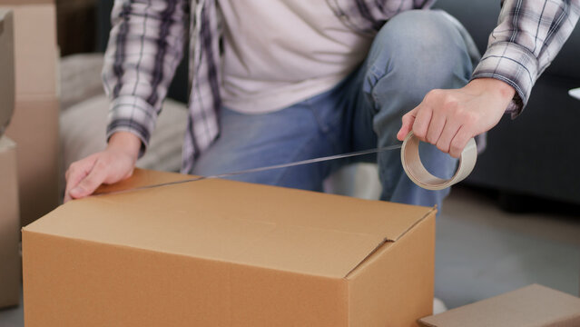 Young Asian man sealing cardboard box with packing tape during home move in modern living room