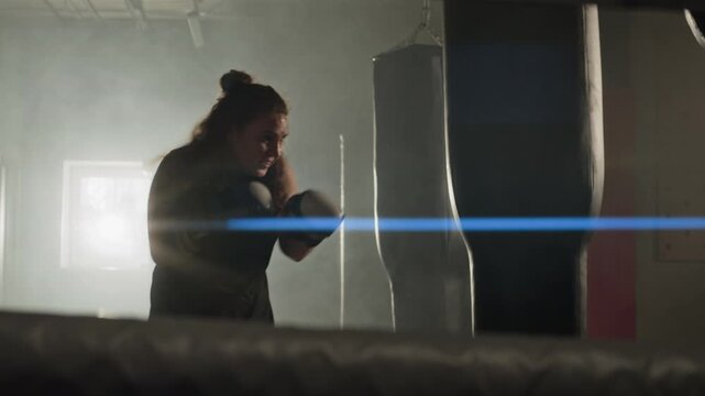 Young Caucasian woman wearing boxing gloves training and punching heavy bag in boxing gym. Perfect for fitness brand advertisements, gym promotions, and motivational campaigns