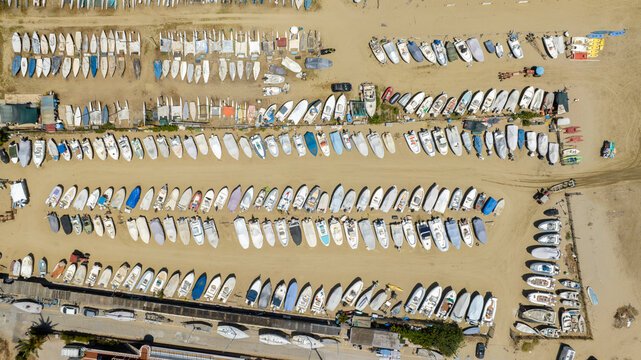 Top-down aerial view of many small boats and dinghies neatly parked in parallel rows on a sandy beach storage area, showing an organized pattern near the sea shore during a sunny day. Zenithal shot.