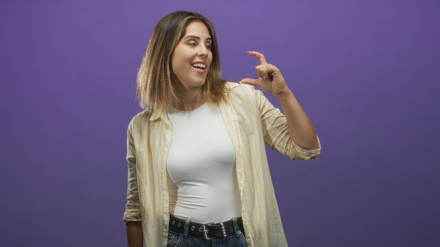 Woman with blonde hair and striped shirt pinches fingers to indicate tiny size while wearing white top and jeans in studio; playful curiosity.