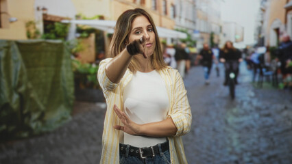 Fototapeta premium Woman pointing finger forward with crossed arm and stern expression on a cobblestone street with cyclists and cafe in view; confidence warning resolve.