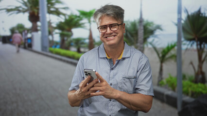 Fototapeta premium Man with grey hair and glasses holding smartphone with both hands and smiling on a seaside promenade with palm trees and ocean in view; contentment ease.