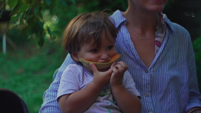 Toddler eating cantaloupe while sitting on mother&rsquo;s lap in a garden, healthy outdoor snacking expressing family connection, comfort and nurturing care