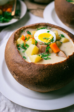 Polish sour rye soup with sausage, potatoes, carrots and a halved egg served in a bread bowl. Comfort food, traditional cuisine, rustic dining, hearty meal, cozy lunch, farmhouse cooking.