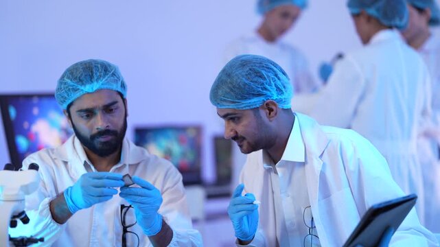 Two masked scientists discuss findings on a tablet as teammates operate a microscope behind them under atmospheric blue light in a biotechnology research facility