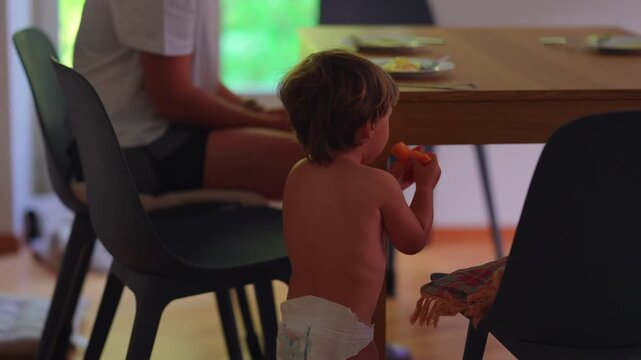 Toddler eating melon slice at dining table in family home, independently enjoying healthy snack, expressing curiosity, childhood development, nutrition, everyday learning