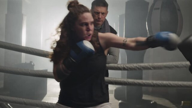 Two Caucasian young women sparring in boxing ring while middle aged Caucasian trainer watches. Footage ideal for fitness brands, sports advertisements, and motivational gym campaigns