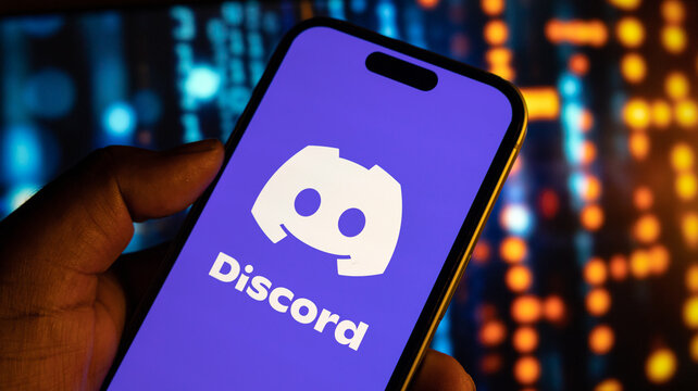 A close up of a hand holding an iphone with discord logo on screen against blurred background of binary code.
