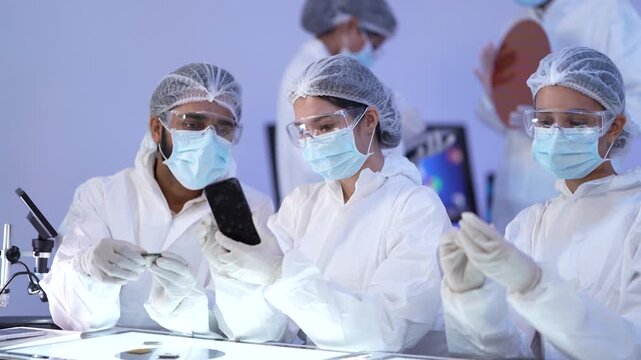 Cleanroom team inspects a small dark specimen held up for close examination among masked colleagues in a sterile controlled biotechnology research laboratory setting