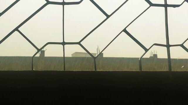 Raindrops falling steadily in bright daylight viewed through a dark decorative metal window grill against a hazy outdoor landscape background during monsoon