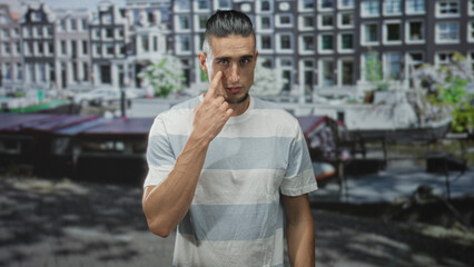 Fototapeta premium Man pointing finger to eye and touching cheek in front of canal building, standing and looking intently at camera; contemplation.