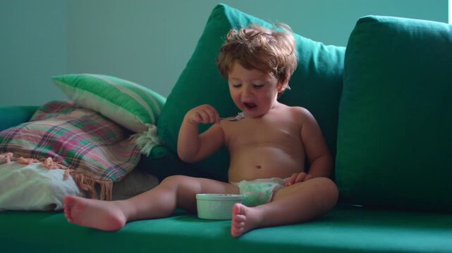 Toddler eating yogurt independently on sofa at home, enjoying healthy snack, expressing self feeding, childhood independence, nutrition, everyday family life and early development