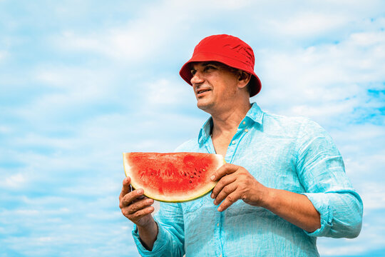 Mature man in red hat and blue shirt holds juicy slice of watermelon while relaxing outdoors on a bright summer day. Concept of summer relaxation, consuming organic, juicy fruit while thirsty
