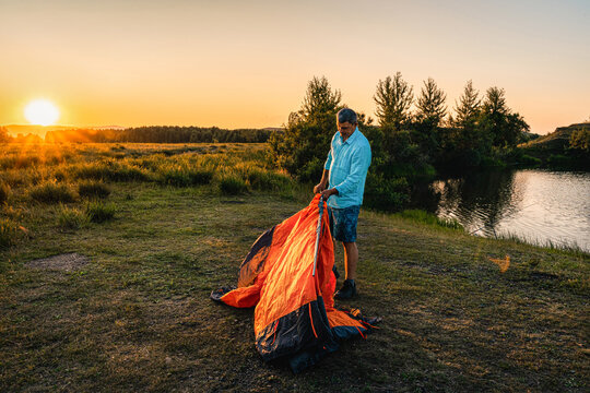 Man pitches an orange tent by calm river during golden hour, enjoying freedom, adventure, nature during his summer vacation. Intentional Solitude