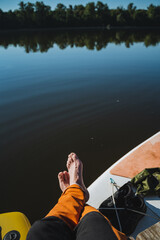 Fototapeta premium Person relaxing on paddleboard with bare feet over calm lake, reflected trees, orange pants, shoes and rope nearby, serene outdoor leisure scene under clear sky