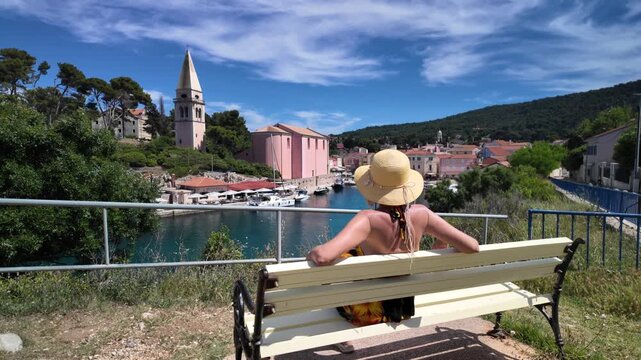 Woman wearing straw hat relaxing on bench, looking at charming harbor of Mali Losinj, Croatia with colorful buildings, church bell tower,and lush green hills under blue sky of Losinj Island in Croatia