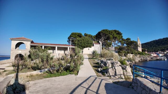 Stone cemetery chapel featuring a red wooden door and ornate ironwork, positioned on a hill overlooking the deep blue Adriatic Sea under a clear summer sky on Losinj Island in Croatia