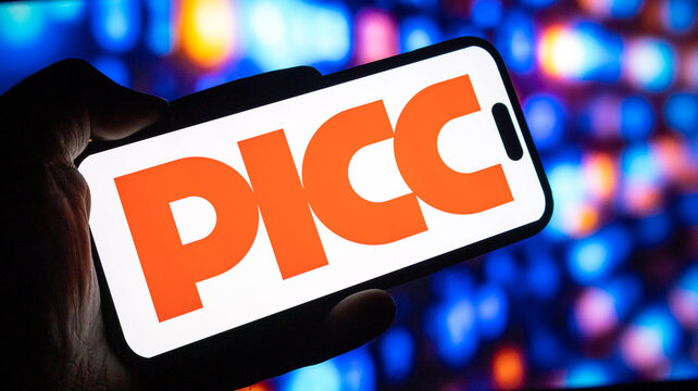 Picc insurance company logo displayed on smartphone held in hand against blurred digital lights on monitor screen display.