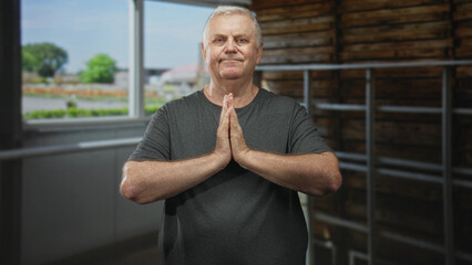 Fototapeta premium Senior man stands with palms pressed together in prayer gesture in a studio by a window and wooden wall, wearing a grey tshirt; calm reflection.