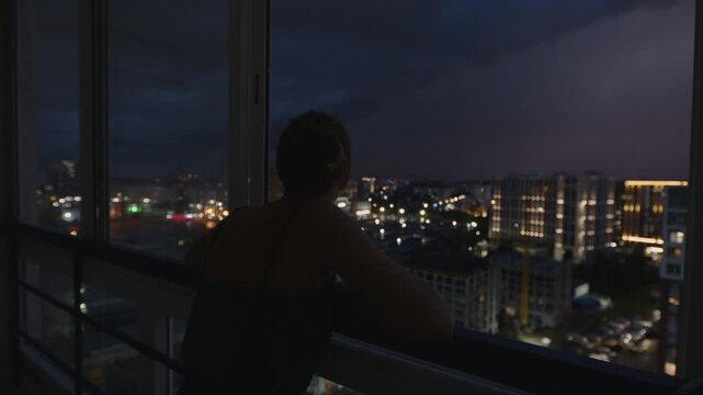 Storm over the city. Woman watches thunder storm with lightnings from the balcony of her apartment on a top floor with city view on the background
