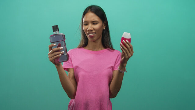 Young thai woman holding a mouthwash bottle and small toothpaste tube, scrunching face in a studio; hesitation decision disgust.