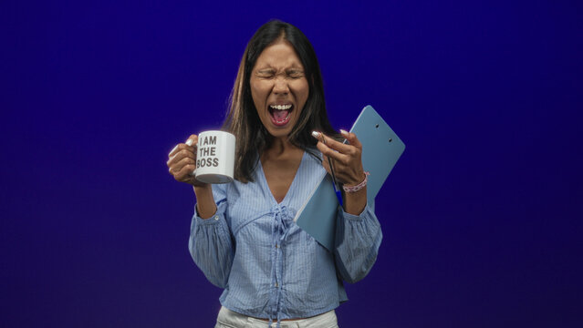 Young thai woman holds mug reading i am the boss and blue clipboard, mouth open and eyes closed while screaming in studio; leadership frustration.