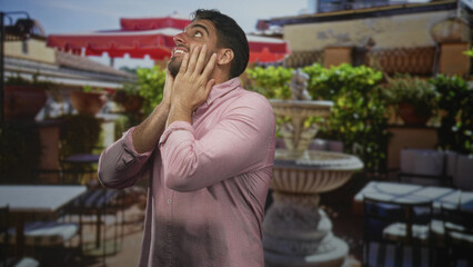 Fototapeta premium Man with hands covering cheeks looking upward on building terrace beside stone fountain, wearing pink shirt; surprise contemplation.