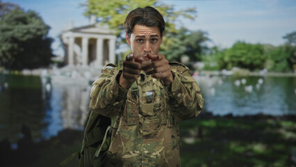 Fototapeta premium Man soldier in camouflage with hands clasped then open palms and pointing, tongue out, wearing dogtags and backpack near a classical building by a lake; playful camaraderie.