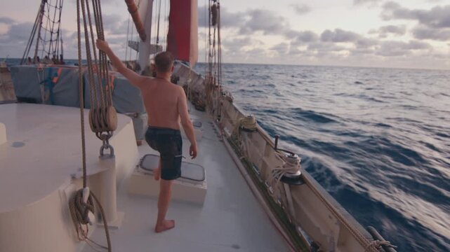 Sailing the Atlantic. Male sailor stands on the deck of the big sailing boat moving in the Atlantic Ocean under the sails and enjoys the first light of the day