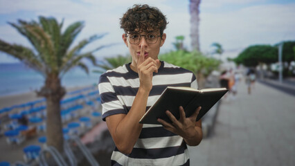 Fototapeta premium Teen boy in striped shirt and round glasses holds an open notebook and places finger to lips in a shushing gesture on a palm lined street by the beach; quiet mischief.