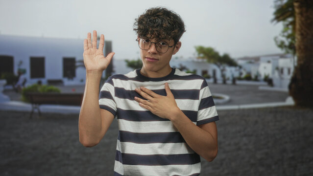 Teen boy with glasses places hand on chest and raises right hand in an oath gesture on a street lined with white buildings and a bench; sincere promise.