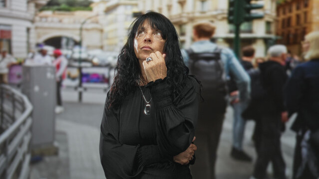 Senior woman touching chin with hand on a busy urban street crossing by trafficlight and crowds while gazing upward; contemplation.