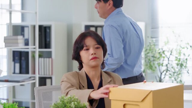 A somber businesswoman packs a photo frame into a cardboard box at her desk while a colleague clears out background items, symbolizing job loss and workforce displacement caused by AI automation.