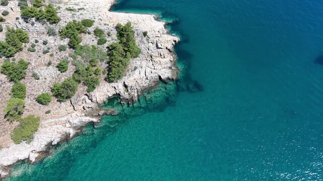 Oblique aerial view drifting along a pale rocky shoreline bordered by transparent turquoise shallows and deep blue sea.