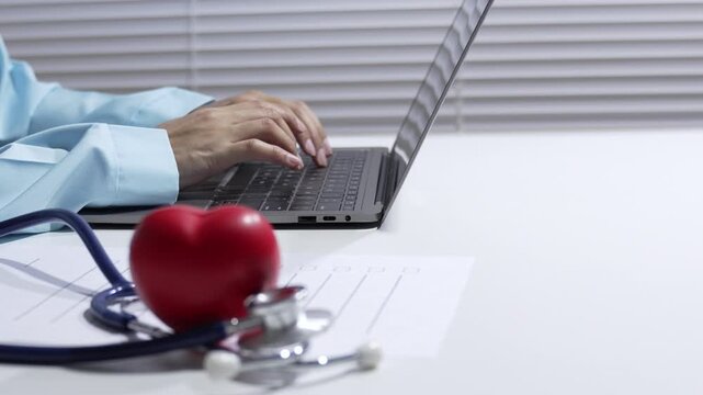Doctor typing on laptop with stethoscope and red heart on desk in medical office, telemedicine healthcare consultation, medical data entry, remote care