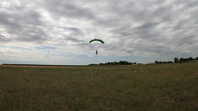 A skydiver with a blue-green parachute landing in an open field amidst a picturesque setting