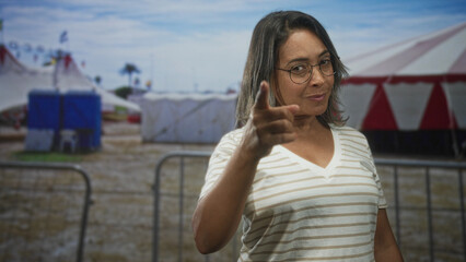 Obraz premium Hispanic middle aged woman points finger to camera near red and white circus tents in street carnival setting; confidence invitation.