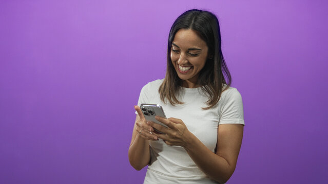 Woman tapping smartphone with finger in studio against purple backdrop, smiling while reading messages, wearing white t shirt; joy.