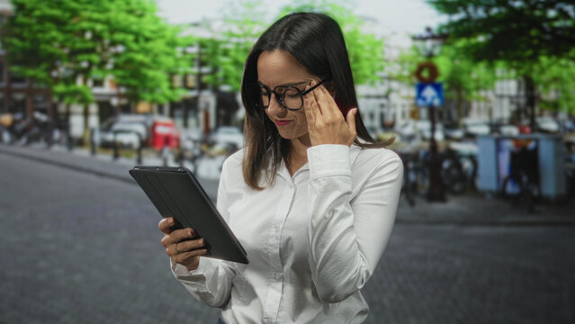 Woman in white shirt holding a tablet with one hand and touching glasses at her temple while reading on a street; concentration study.