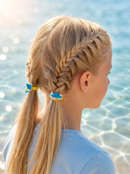 Girl with blonde hair styled in two French braids decorated with cute cartoon fish hair elastic accessories. Summer seaside vacation hairstyle for children. Fun sea animal accessory.