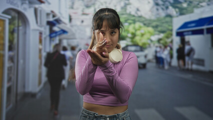 Fototapeta premium Young woman points finger forward in a finger gun gesture with white headphones around neck and cropped top revealing midriff on a busy street; confident attitude.