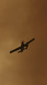 Silhouette of a firefighting aircraft flying low through dense, sepia toned smoke from a forest fire. Emergency response to a natural disaster