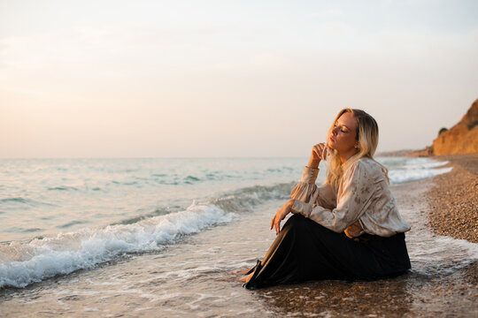 Lonely young woman wearing office clothes sitting in sea water at coast line outdoors. Loneliness.