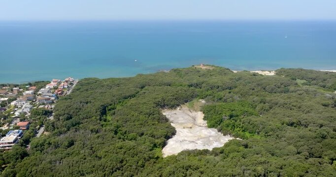 Aerial high-angle view showing a natural geothermal solfatara area nestled within a lush green forest, situated along a scenic coastline with deep blue mediterranean sea in a sunny summer morning.