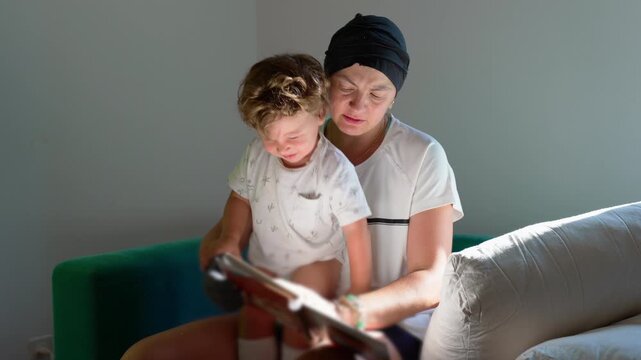 Mother recovering from cancer reads an illustrated story to her toddler son on the sofa at home, sharing hope, resilience, learning and loving family connection
