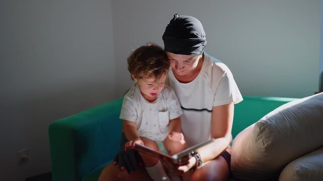 Mother reads an illustrated book with her toddler son seated on her lap on the sofa at home, promoting early literacy, focused education and close family connection