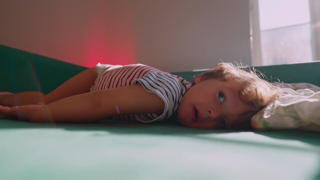 Pensive toddler resting on sofa beside a pillow at home, quiet childhood pause expressing fatigue, contemplation, vulnerability and emotional wellbeing