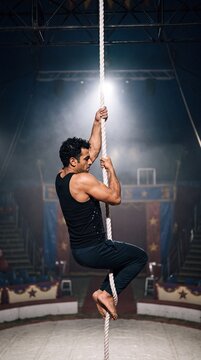 Muskul&ouml;ser Zirkusartist klettert &uuml;ber die Schulter gesehen an einem wei&szlig;en Vertikalseil in der Manege im Scheinwerferlicht