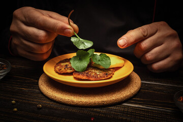 A chef arranges fresh herbs on top of patties placed on a yellow plate. The setting is a restaurant kitchen with dim lighting in the evening © Віталій Б.