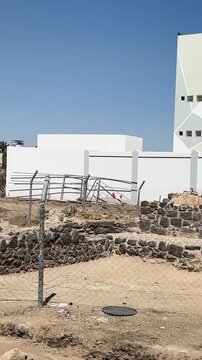 Fenced ancient stone ruins and excavation site next to modern buildings.